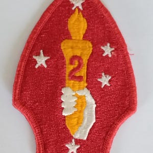 Op de afbeelding: Een rood geborduurd embleem met een gele kaars met het rode nummer 2. Vier witte sterren omringen de kaars. De patch heeft een puntige bovenkant en een gebogen onderkant.