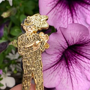 May include: Gold-colored enamel pin featuring a cartoon character in a pinstripe suit and fedora, holding a gun and a briefcase. The character is smoking and the speech bubble above their head reads "3 for 20 deals".
