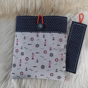 Könnte beinhalten: Ein nautisch thematisches Buchcover- und Lesezeichen-Set. Das Buchcover hat einen hellgrauen Stoff mit einem Muster aus Ankern, Leuchttürmen und Rettungsringen. Die Oberseite ist marineblau mit weißen Punkten. Das Lesezeichen hat ein marineblaues Pünktchenmuster und eine rote Schlaufe.