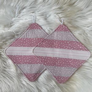 Könnte beinhalten: Zwei rosa und weiße, gesteppte Topflappen in Diamantform. Sie haben einen rosa Rand, einen weiß gestreiften Abschnitt und einen rosa gepunkteten Abschnitt. Jeder Topflappen hat eine weiße Schlaufe zum Aufhängen.
