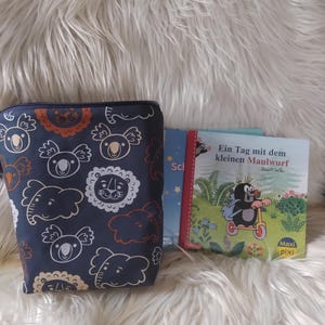 Könnte beinhalten: Eine marineblaue Reißverschlusstasche mit einem sich wiederholenden Muster aus Koala- und Löwenillustrationen. Zwei Kinderbücher sind zu sehen, eines mit dem Titel "Ein Tag mit dem kleinen Maulwurf". Die Artikel liegen auf einer flauschigen weißen Oberfläche.