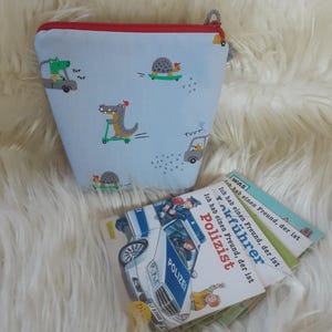 Könnte beinhalten: Hellblaue Reißverschlusstasche mit rotem Rand, mit einem verspielten Aufdruck von Cartoon-Alligatoren und Schildkröten auf Rollern und Autos. Mehrere kleine Bücher mit deutschem Text und einer Polizei-Auto-Illustration liegen daneben.