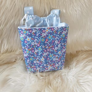 Könnte beinhalten: Eine kleine Tasche mit floralem Muster auf blauem Grund. Die Tasche hat ein hellblaues Innenfutter und einen Kordelzugverschluss. Zwei hellblaue Träger mit weißen Punkten sind oben befestigt. Die Tasche ist mit bunten Blumen verziert.