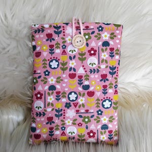 Könnte beinhalten: Eine rosa Stoffhülle mit Blumenmuster und Knopfverschluss. Das Muster wiederholt stilisierte Blumen in Weiß, Gelb, Rot und Blau. Ein Holzknopf mit dem Text "Handmade with love" sichert den Verschluss. Die Hülle ist wahrscheinlich für ein Tablet oder einen E-Reader.