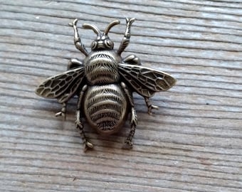May include: A detailed bronze-colored bee brooch with outstretched wings, antennae, and intricate detailing. The bee is positioned on a weathered, light brown wooden surface, showcasing the insect's textured body and delicate wings.