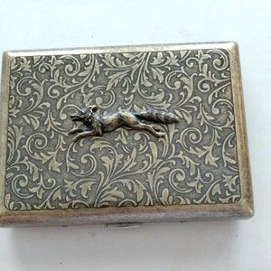 May include: Silver-toned rectangular case with an embossed floral pattern and a running fox emblem. The case has a hinged lid and a metallic finish, suggesting it may be a cigarette case or similar item.