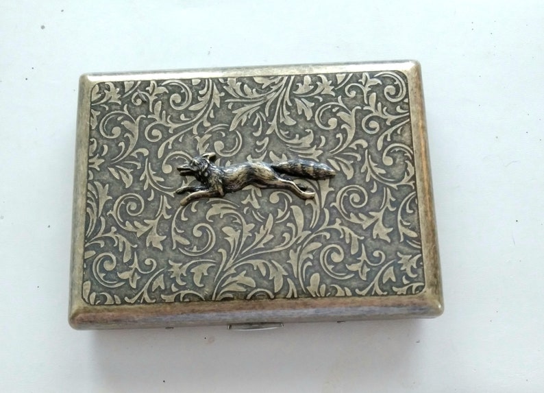 May include: Silver-toned rectangular case with an embossed floral pattern and a running fox emblem. The case has a hinged lid and a metallic finish, suggesting it may be a cigarette case or similar item.