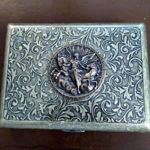 Pode incluir: Caixa met&aacute;lica retangular com um padr&atilde;o floral ornamentado e rodopiante. Um medalh&atilde;o circular com uma figura alada montando um cavalo est&aacute; centrado na tampa. A caixa tem um esquema de cores bronze e prata.