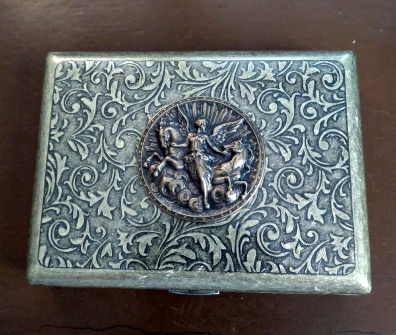 Pode incluir: Caixa met&aacute;lica retangular com um padr&atilde;o floral ornamentado e rodopiante. Um medalh&atilde;o circular com uma figura alada montando um cavalo est&aacute; centrado na tampa. A caixa tem um esquema de cores bronze e prata.