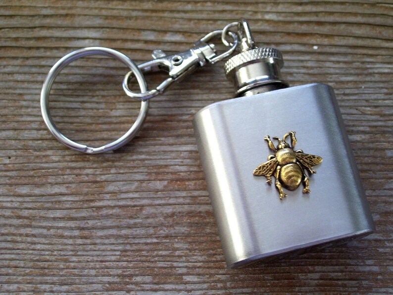 May include: A stainless steel flask with a silver keychain and a decorative brass bee. The flask has a screw-on cap and a rectangular shape. The bee is centered on the front of the flask.