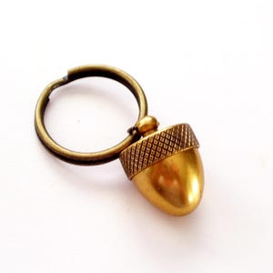 May include: A gold-toned acorn-shaped keychain with a ring attached. The acorn has a textured top and a smooth, rounded bottom.