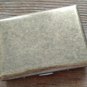 May include: A rectangular, metallic cigarette case with a textured, brushed finish. The case is a muted gold color and has rounded corners. The case is closed and sits on a wooden surface.