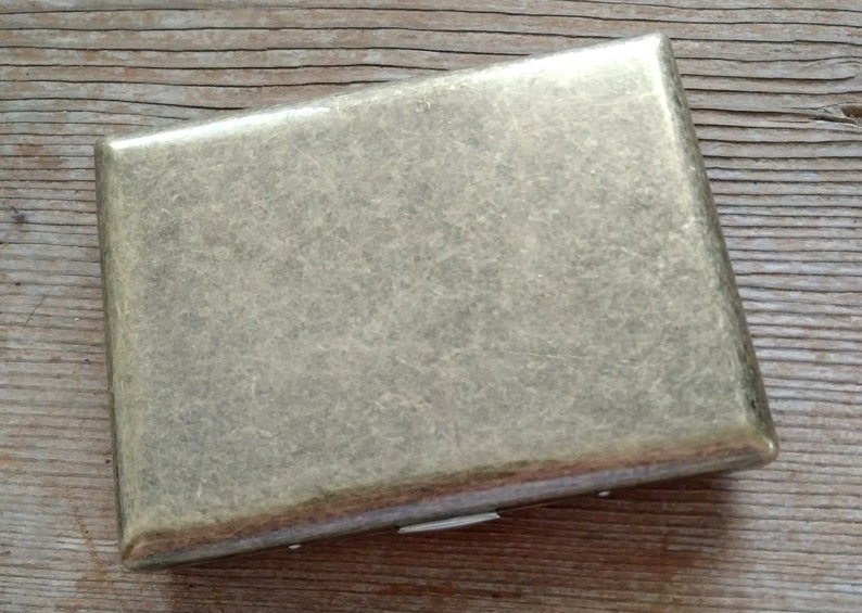 May include: A rectangular, metallic cigarette case with a textured, brushed finish. The case is a muted gold color and has rounded corners. The case is closed and sits on a wooden surface.