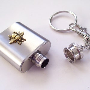 May include: A silver flask with a decorative brass bee emblem. The flask has a screw-on cap attached to a keychain. The flask is rectangular and has a brushed metal finish.