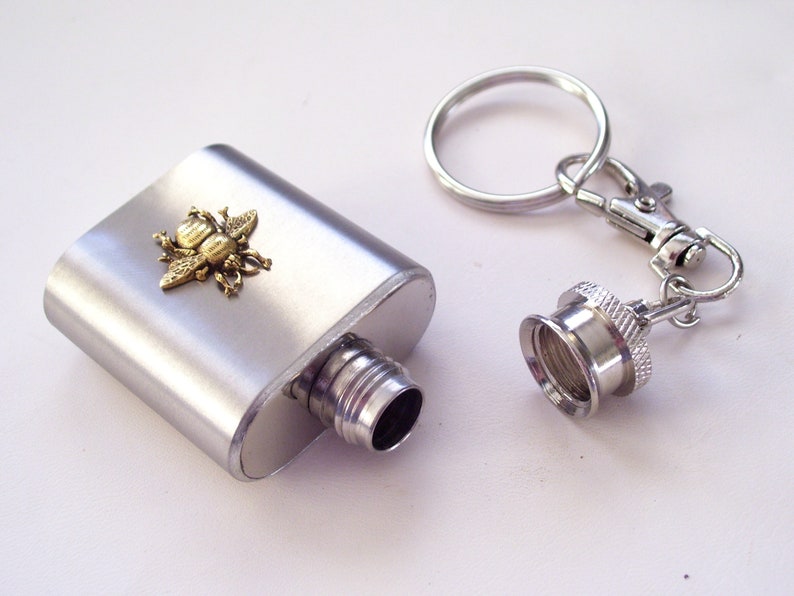 May include: A silver flask with a decorative brass bee emblem. The flask has a screw-on cap attached to a keychain. The flask is rectangular and has a brushed metal finish.