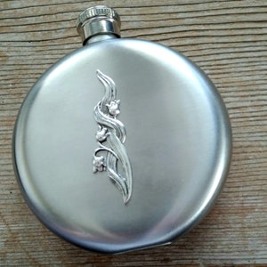 May include: A round, brushed stainless steel flask with a silver floral design. The flask has a screw-on cap and is set against a wooden background. The floral design is an elegant, stylized depiction of lily of the valley.