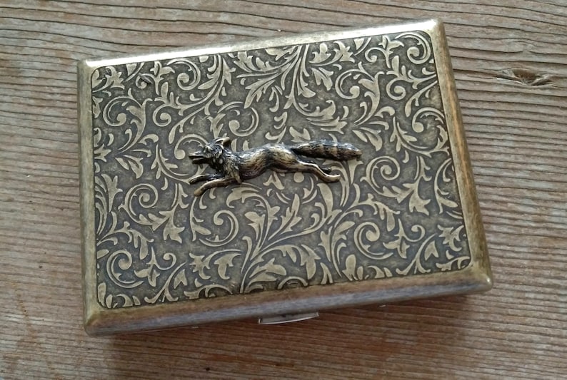 May include: A rectangular, antique-style cigarette case with a detailed floral pattern and a running fox emblem. The case has a metallic finish with a hinged closure, and is likely made of metal.