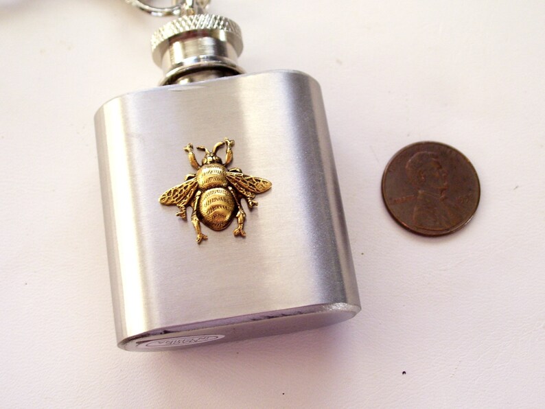May include: A stainless steel flask with a silver cap and a decorative gold-colored bee emblem. The flask is rectangular and has a brushed metal finish. A penny is next to the flask for size comparison.