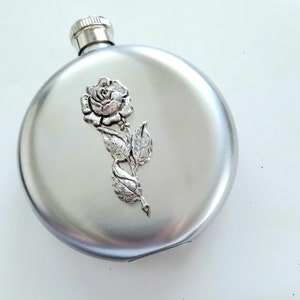 May include: A silver-colored flask with a decorative raised rose and leaf design. The flask has a rounded shape with a screw-on cap. The rose is detailed with petals and leaves.