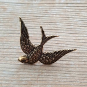 May include: A bronze-colored swallow bird pin with outstretched wings. The bird is detailed with etched feathers and a small, round head. The pin is set against a light brown wooden surface.