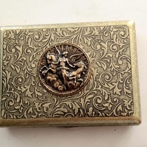 Pode incluir: Estojo de metal retangular com um padr&atilde;o ornamental e rodopiante e um medalh&atilde;o central com uma figura alada montada em dois cavalos. O estojo tem um esquema de cores bronze e prata.