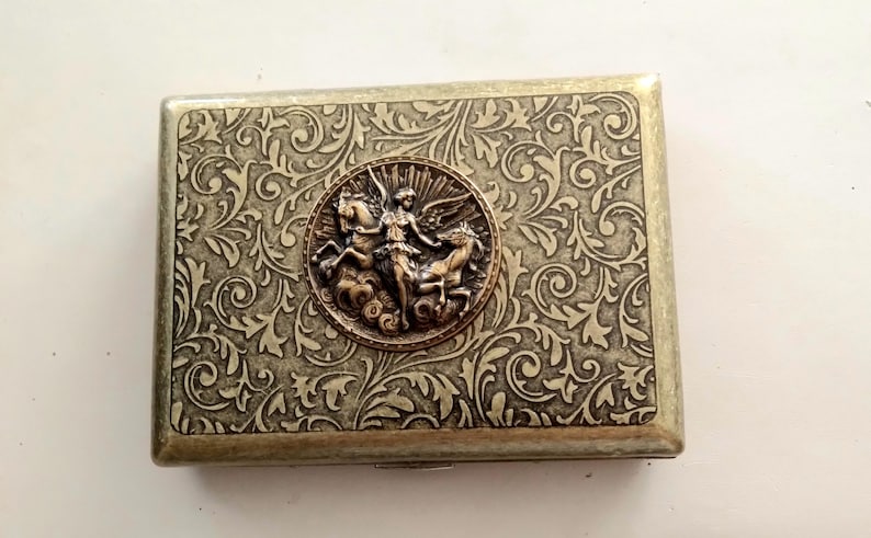 Pode incluir: Estojo de metal retangular com um padr&atilde;o ornamental e rodopiante e um medalh&atilde;o central com uma figura alada montada em dois cavalos. O estojo tem um esquema de cores bronze e prata.