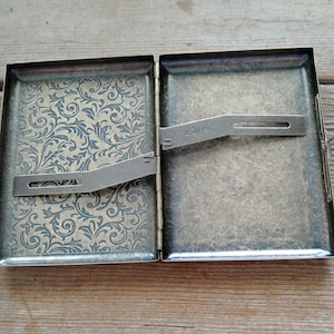 May include: Open antique brass cigarette case. The interior features a floral pattern on one side and a metal clip. The case is hinged and has a clasp closure. The word "Guipa" is stamped on the metal clip.