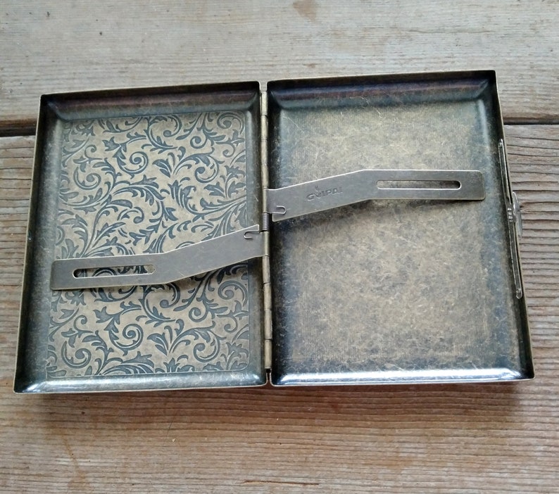 May include: Open antique brass cigarette case. The interior features a floral pattern on one side and a metal clip. The case is hinged and has a clasp closure. The word "Guipa" is stamped on the metal clip.