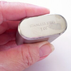 May include: A stainless steel flask with a silver finish. The text "STAINLESS STEEL" and "1 oz" are printed on the flask. The flask is oval-shaped and held in a hand.