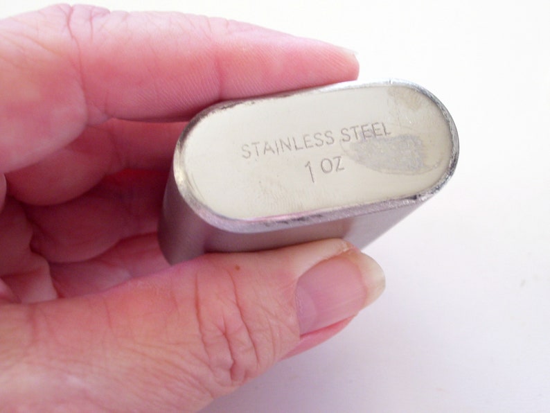 May include: A stainless steel flask with a silver finish. The text "STAINLESS STEEL" and "1 oz" are printed on the flask. The flask is oval-shaped and held in a hand.