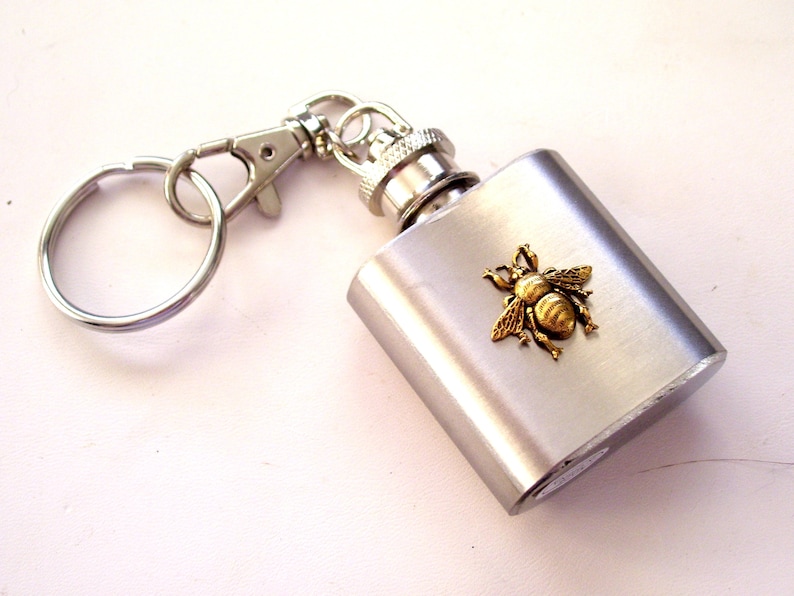 May include: A silver-colored flask with a gold-colored bee emblem. The flask has a screw-on cap and a keychain attachment. The bee is detailed with textured wings and body. The flask is rectangular and has a brushed metal finish.