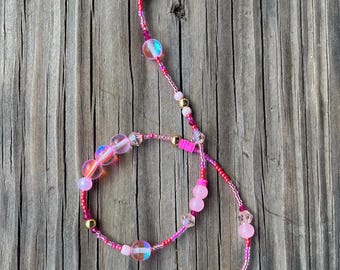 May include: Two pink beaded necklaces with iridescent beads and gold accents. The necklaces are layered on a wooden surface.
