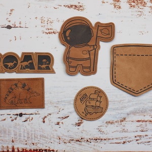 Könnte beinhalten: Fünf braune Lederpatches mit verschiedenen Designs. Die Patches beinhalten einen Dinosaurier, einen Astronauten, ein Schiff, eine Tasche und das Wort "ROAR".