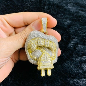 Op de afbeelding: Een gouden hanger in de vorm van een mond die in een stekker bijt, volledig bezet met heldere en gele zirkonia-stenen.