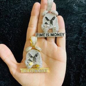 Könnte beinhalten: Zwei diamantbesetzte Anhänger in Form von Weckern, einer silberfarben und einer goldfarben. Jeder Anhänger hat ein schwarzes Zifferblatt und darunter die Worte "TIME IS MONEY". Die Anhänger werden auf einer Hand präsentiert.