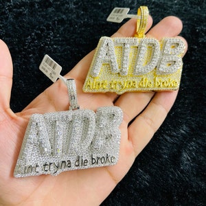May include: Two gold pendants with the letters "A.T.D.B." set with clear cubic zirconia stones. The pendants are on a chain and have the text "Ain't tryna die broke" engraved on the bottom.