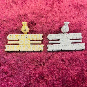 May include: Two gold and silver pendants with the words "Get Rich Or Die Tryin" and a money bag design. The pendants are on a red velvet background.