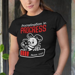 Könnte beinhalten: Ein schwarzes T-Shirt mit einem weißen Comic-Kaninchen, das ein Telefon und eine Tasse Kaffee hält. Der Text "Procrastination in PROGRESS" ist in Rot über dem Kaninchen gedruckt. Unter dem Kaninchen befindet sich ein Batteriesymbol mit drei roten Balken und dem Text "Bitte warten".