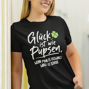 Peut inclure: Un t-shirt noir avec un motif blanc qui dit "Glück ist wie pupsen. Wenn man es erzwingt wird es scheiße." Le motif comprend également un trèfle à quatre feuilles vert.
