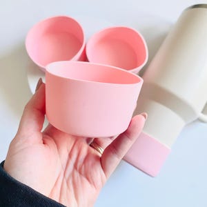 May include: Three pink silicone cup lids, one in a hand, two on a white surface. The lids are designed to fit on top of cups to prevent spills.
