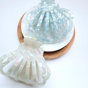 May include: Two shell-shaped hair claws with a glossy finish. One claw features a light blue iridescent mosaic pattern, while the other has a pearlescent white marbled appearance. Both accessories have a curved, multi-pronged design.