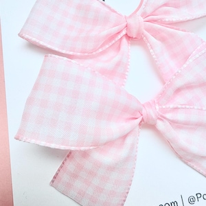 May include: Two pink and white gingham hair bows. The bows are tied in the center and have a classic bow shape. The bows are on a white card with the text "www.Pampanitas.Etsy.com | @Pa".
