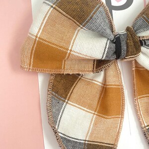 May include: Close-up of a plaid hair bow in shades of brown, white, and gray. The bow has a stitched edge and is tied in the center. The bow is on a white card with a pink background.