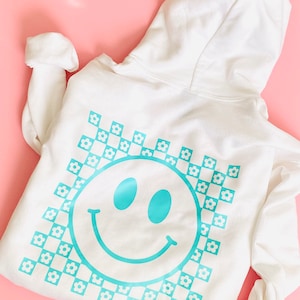 May include: White hoodie with a blue smiley face graphic on the back. The smiley face is surrounded by a checkered pattern with small white flowers.