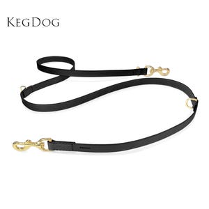 Puede incluir: Una correa para perro de cuero negro con herrajes de metal en tono dorado, que incluye dos ganchos giratorios y dos anillos en D para ajustar la longitud. El nombre de la marca KEGDOG está grabado cerca de uno de los clips, y el texto KEGDOG aparece arriba.