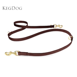 Puede incluir: Una correa para perro de cuero marrón oscuro con herrajes de metal en tono dorado, que incluye dos ganchos giratorios y dos anillos en D para ajustar la longitud. El texto KEGDOG está impreso en negro en la esquina superior izquierda.