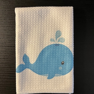 Könnte beinhalten: Weißes Handtuch mit einem blauen Cartoon-Wal-Design. Der Wal lächelt und hat einen Wasserspeier. Das Handtuch hat eine strukturierte Oberfläche. Das Design ist auf dem Handtuch zentriert.
