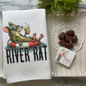 Peut inclure: Serviette de cuisine blanche gaufrée avec un rat de dessin animé portant des lunettes de soleil et une bouée de sauvetage, tenant une bière. Le texte "RIVER RAT" est en noir. L'image comprend également des chocolats et une carte "Thinking of You".