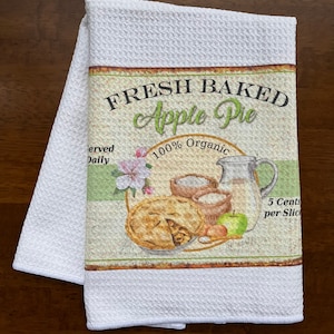 Könnte beinhalten: Weißes Waffel-Küchentuch mit einem Aufdruck von frisch gebackenem Apfelkuchen. Der Aufdruck enthält den Text "FRESH BAKED Apple Pie" und "100% Organic". Außerdem sind eine Kanne, Äpfel und Blumen abgebildet.