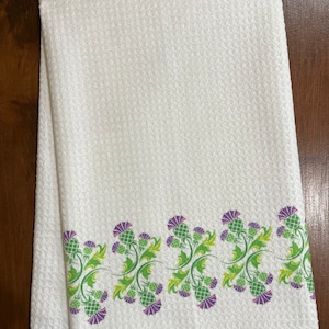 Può includere: Asciugamano da cucina bianco con superficie strutturata e bordo decorativo con motivo floreale di cardi verdi e viola. L'asciugamano è piegato e appoggiato su una superficie di legno.